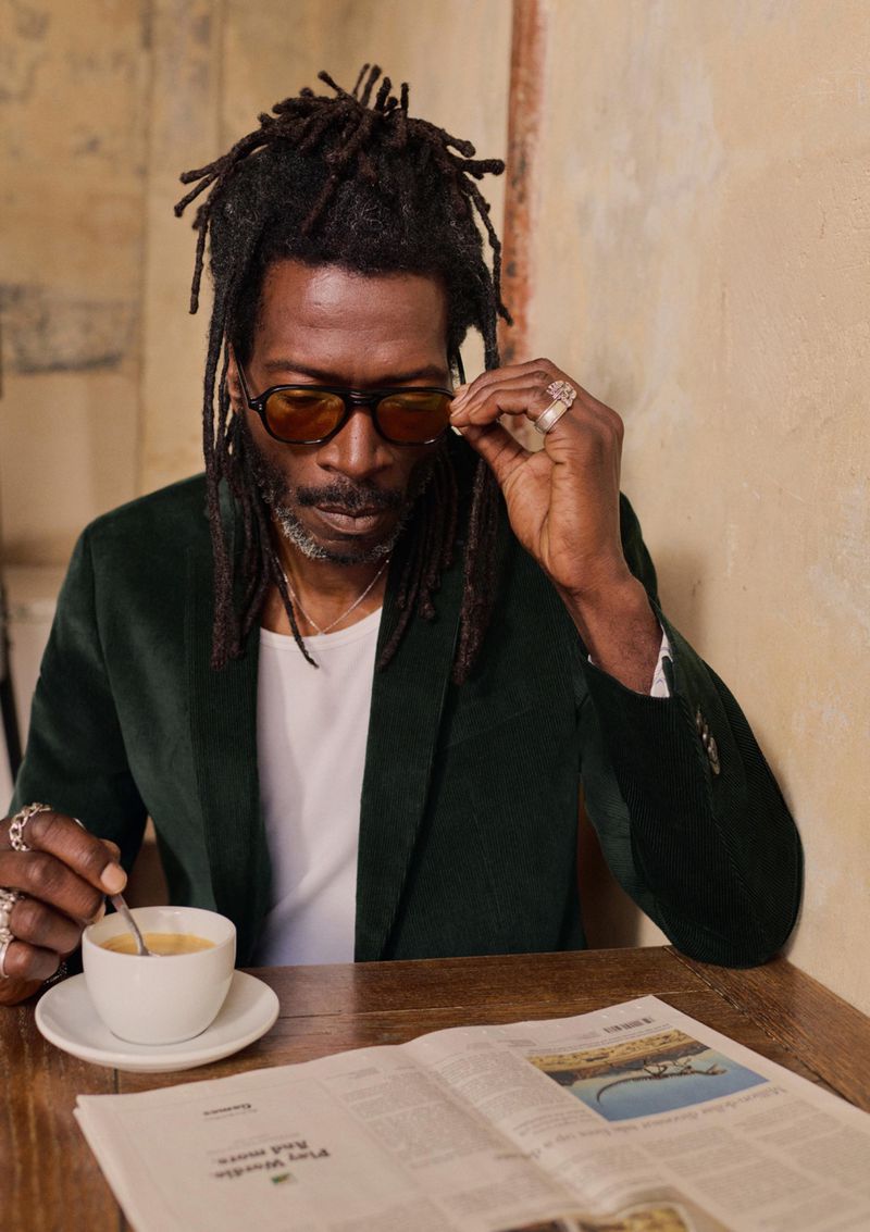 A stylish African-American man with dreadlocks and glasses sips coffee while reading a newspaper at a cafe, wearing a green velvet jacket.