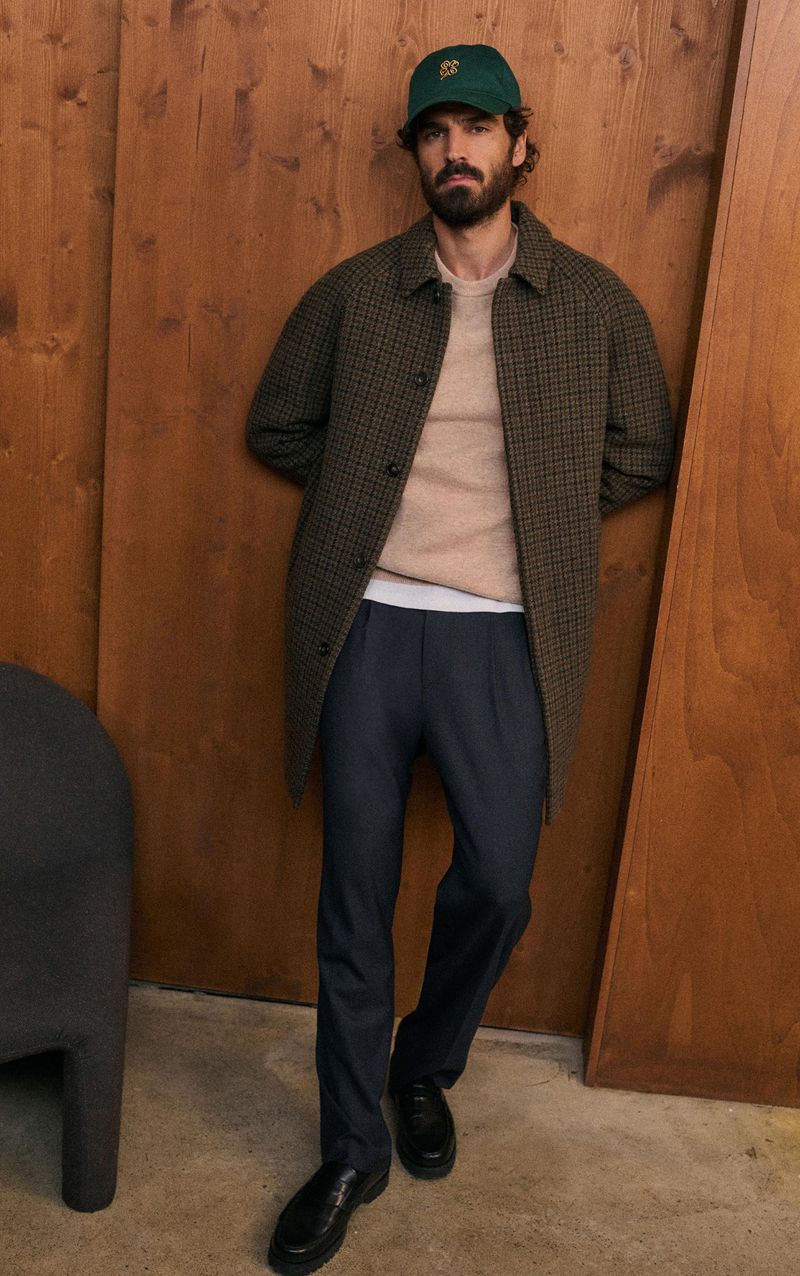 Adult male in a patterned coat, beige sweater, dark pants, black shoes, and a green baseball cap standing against a wooden wall indoors.