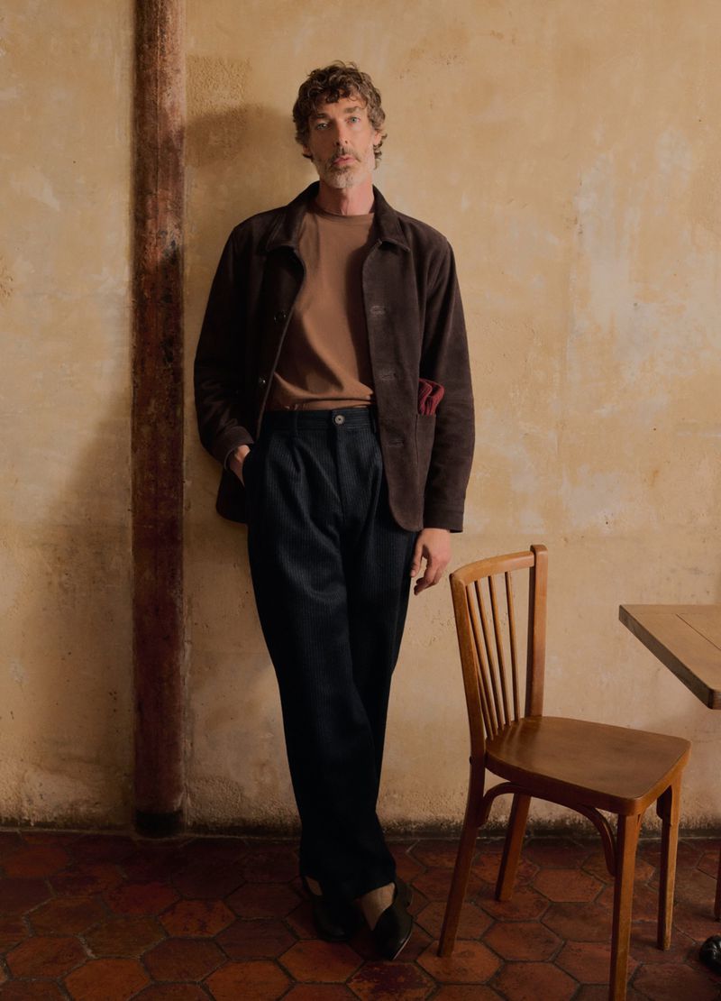 Man wearing a brown jacket standing against a textured wall next to a wooden chair in an editorial portrait