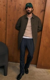 Man wearing a green cap, patterned brown coat, beige sweater, and dark trousers leaning against wood panels.