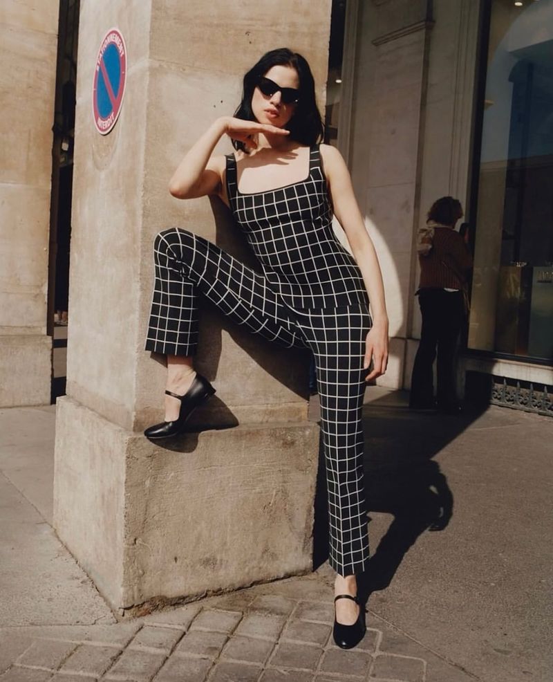 Model Lily McMenamy wearing a black and white grid patterned outfit leaning against a stone pillar outdoors