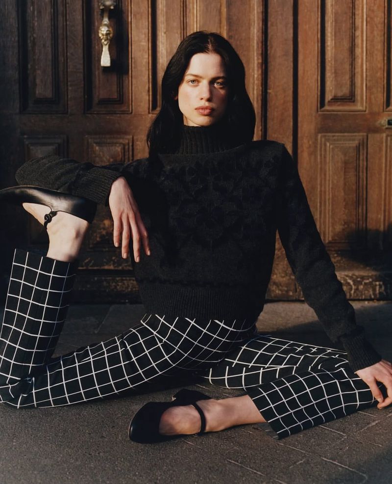 Young woman in black sweater and black and white pants sits on the floor with a wooden door in the background, fashion photography.