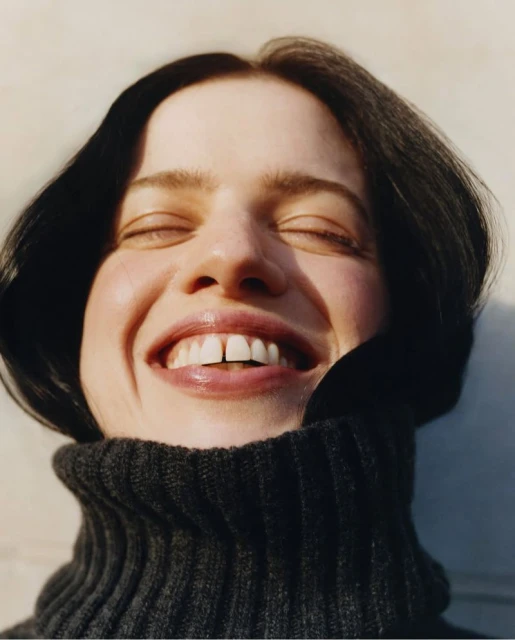 Close-up of a smiling adult woman with closed eyes, wearing a gray turtleneck sweater, against a bright, neutral background in portrait orientation.