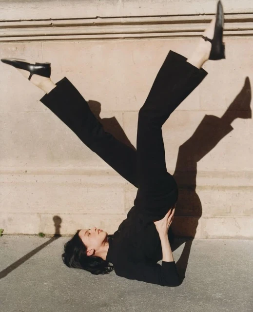 Fashionable woman in black lying on ground with legs up wearing dress shoes, stone wall backdrop and casting stark shadow.