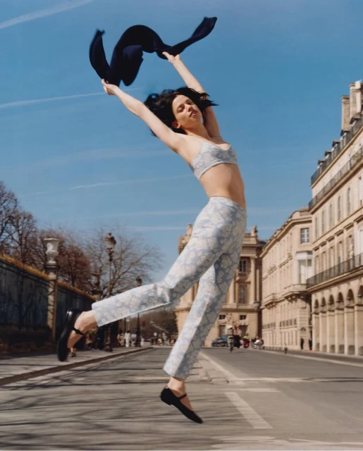 Adult woman leaps into the air while holding a navy scarf on a city street with Parisian architecture and a blue sky.