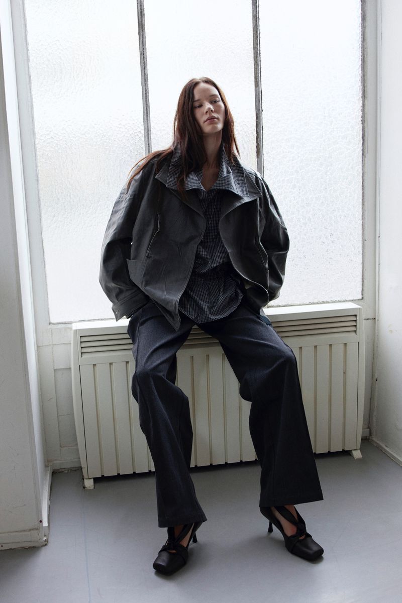 An adult woman with long brown hair sits in front of a window wearing a grey jacket, top, and trousers, along with black heels in an indoor setting.