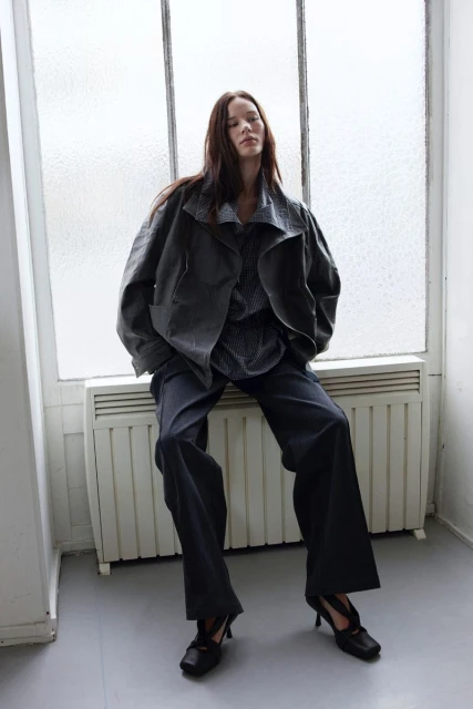 An adult woman with long brown hair sits in front of a window wearing a grey jacket, top, and trousers, along with black heels in an indoor setting.