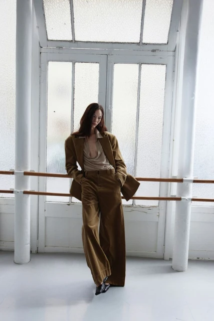 A young woman poses in a tan-colored suit in front of a large window, leaning against a ballet barre in a light-filled room.