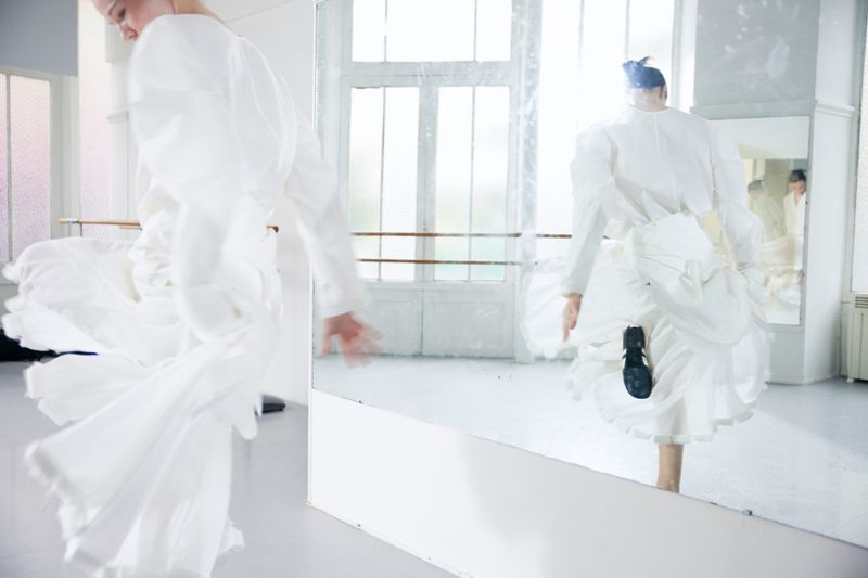 A blurry view of an adult female in a white puffy dress dancing in a bright dance studio, reflected in a mirror with window in background.