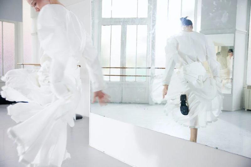 A blurry view of an adult female in a white puffy dress dancing in a bright dance studio, reflected in a mirror with window in background.