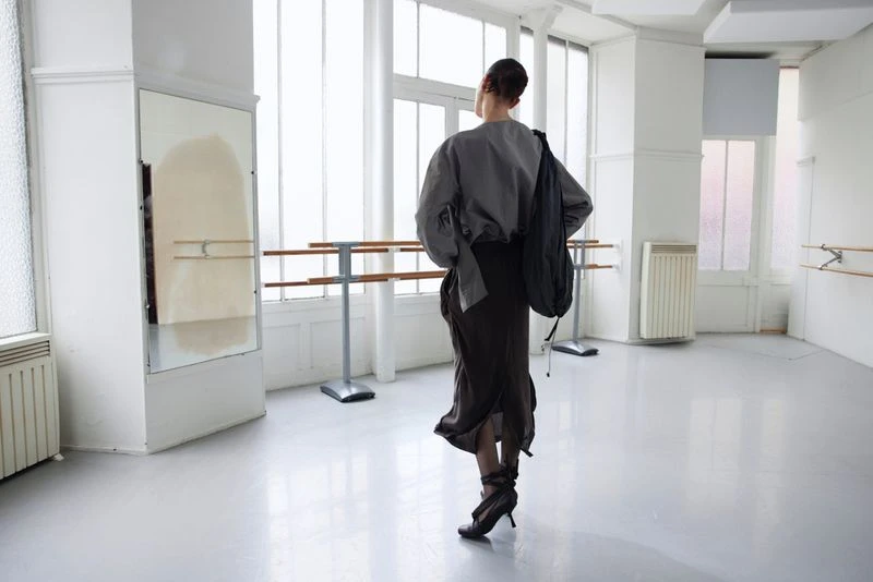 A woman in a gray blouse and dark skirt stands in a bright dance studio, looking away from the camera with a black bag over her shoulder.