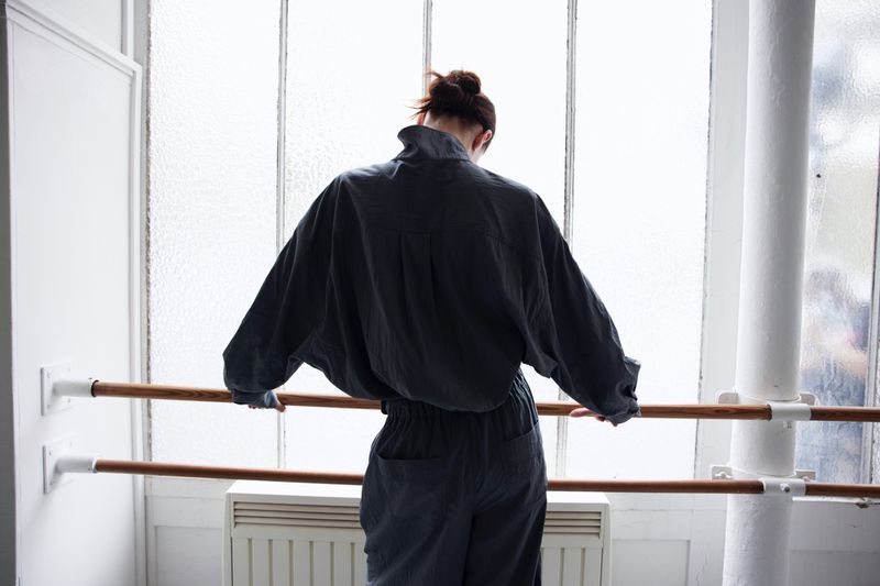 Back view of an adult female in a blue jumpsuit standing in a dance studio, holding onto a ballet barre in front of a window.