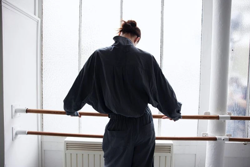 Back view of an adult female in a blue jumpsuit standing in a dance studio, holding onto a ballet barre in front of a window.