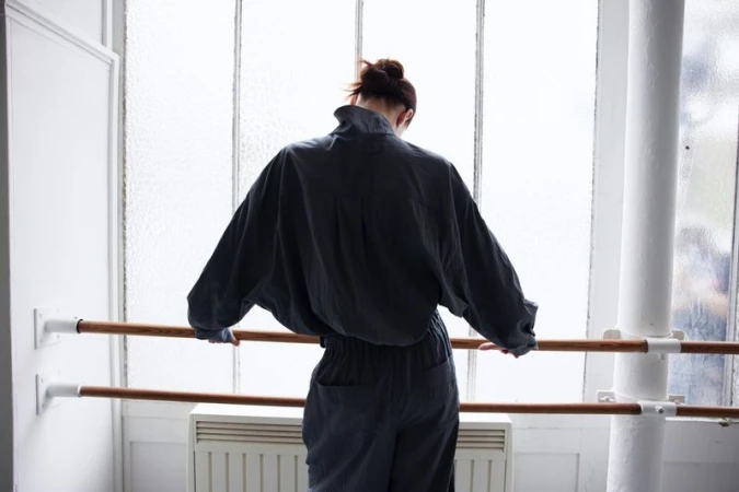 Back view of an adult female in a blue jumpsuit standing in a dance studio, holding onto a ballet barre in front of a window.