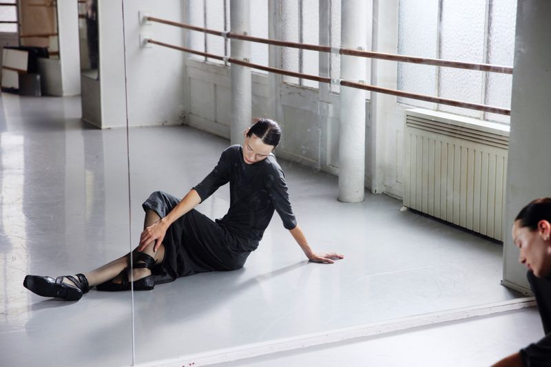 Ballet dancer in black practice clothes rests on the floor of a dance studio with barre and bright light from windows behind.