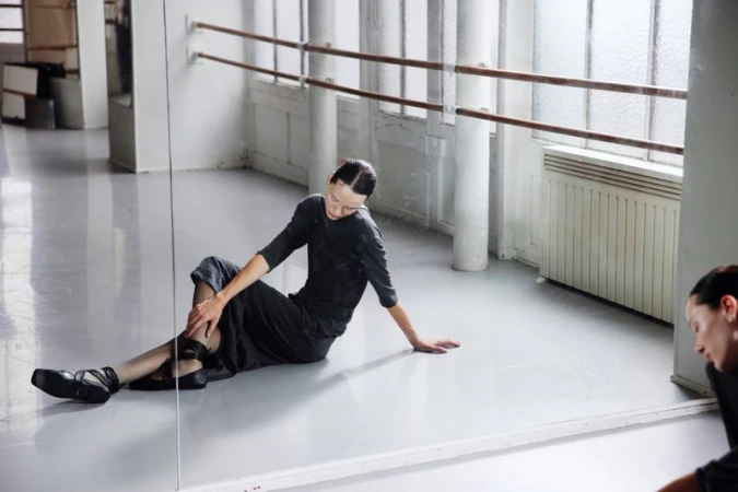 Ballet dancer in black practice clothes rests on the floor of a dance studio with barre and bright light from windows behind.