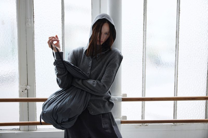 Woman with brown hair wearing a grey hoodie and grey pants standing against a textured-glass window with a wooden ballet bar.