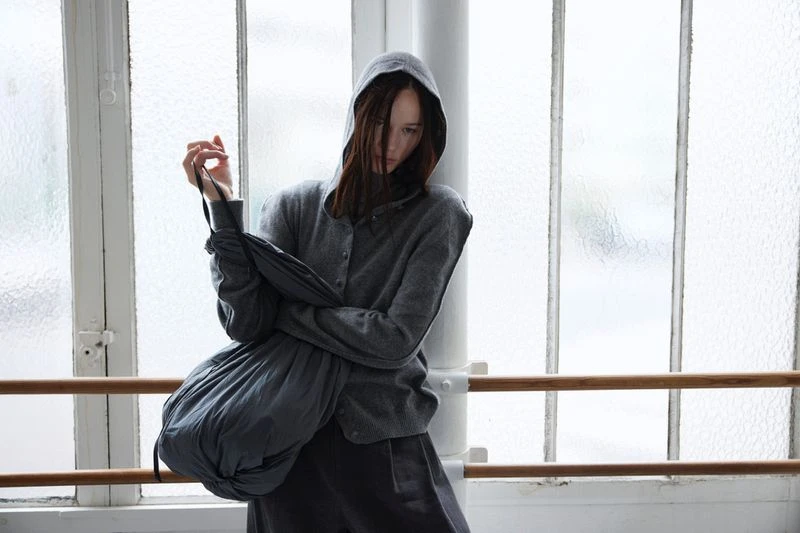 Woman with brown hair wearing a grey hoodie and grey pants standing against a textured-glass window with a wooden ballet bar.