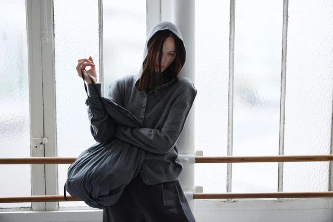 Woman with brown hair wearing a grey hoodie and grey pants standing against a textured-glass window with a wooden ballet bar.