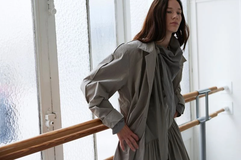 A brunette adult female stands indoors wearing a grey dress and matching coat, posing near a white wall and wooden handrail.