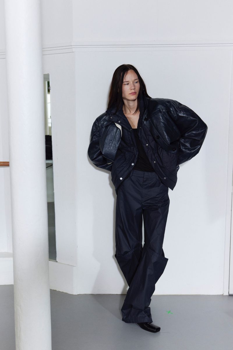 A young adult poses in an indoor studio wearing a black puffer jacket and loose, dark-colored pants against a white background.