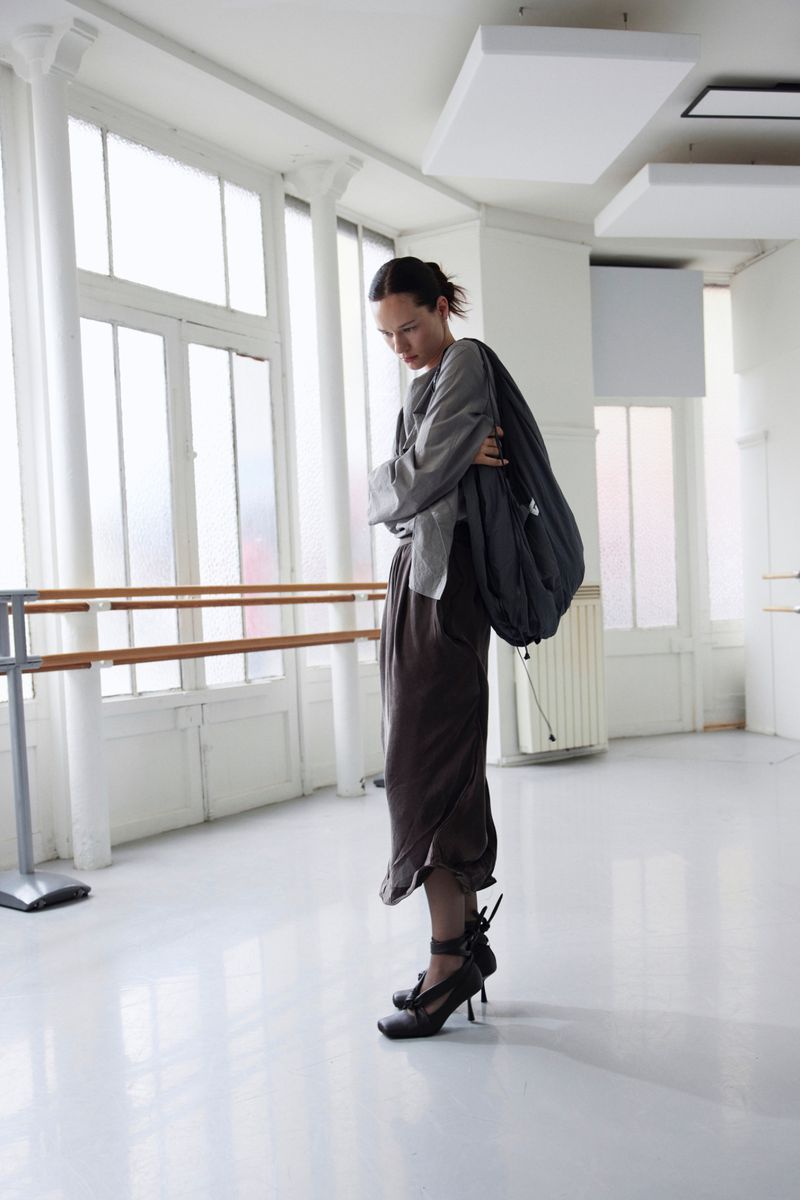 A woman in an outfit stands indoors near a large window with a barre. She is wearing high heels and carrying a large bag.