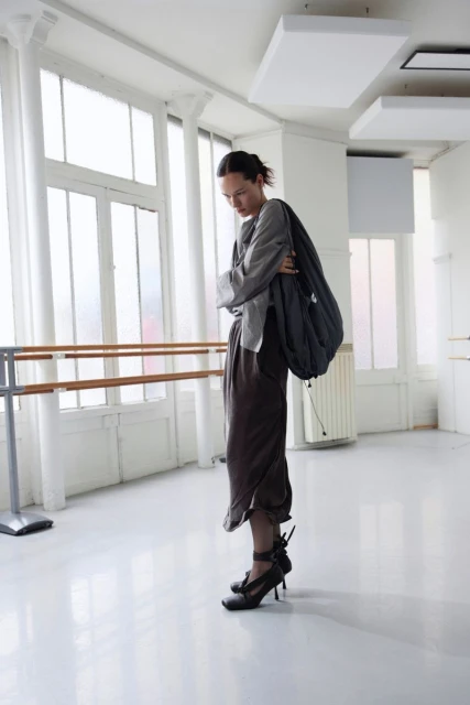 A woman in an outfit stands indoors near a large window with a barre. She is wearing high heels and carrying a large bag.
