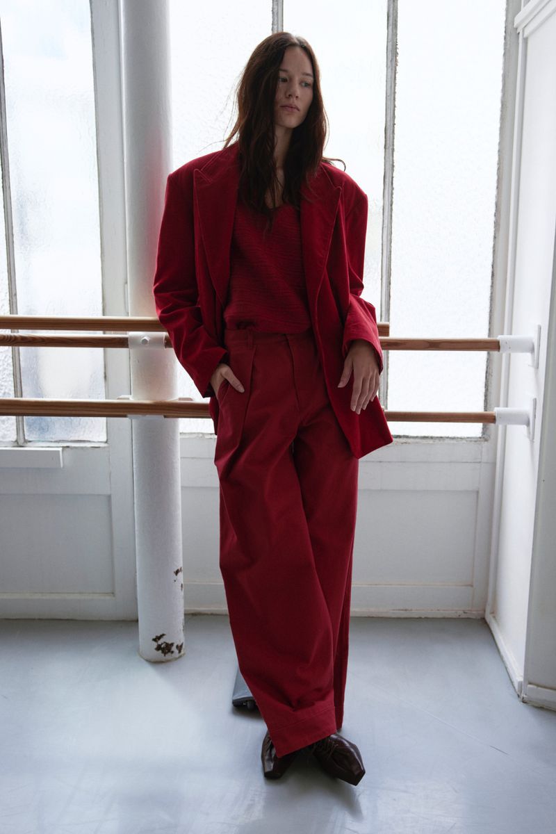 A brunette woman in a striking red suit stands in front of a bright window and white columns, showcasing a bold fashion statement.