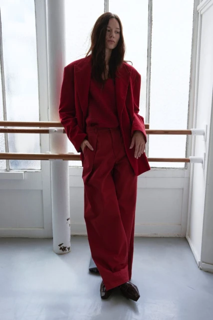 A brunette woman in a striking red suit stands in front of a bright window and white columns, showcasing a bold fashion statement.