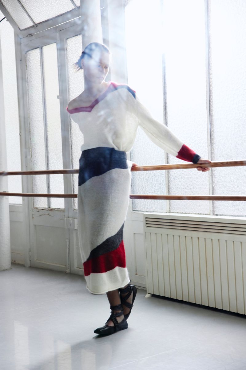 Fashionable woman in white and colorful knitwear posing near ballet barre with large windows, creating a bright, studio setting.