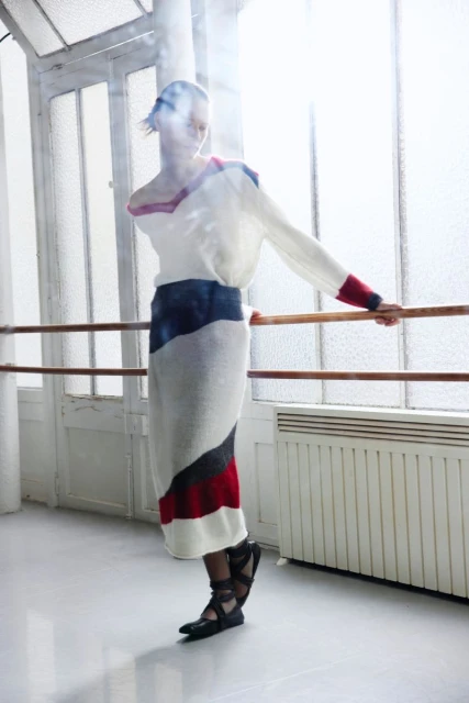 Fashionable woman in white and colorful knitwear posing near ballet barre with large windows, creating a bright, studio setting.