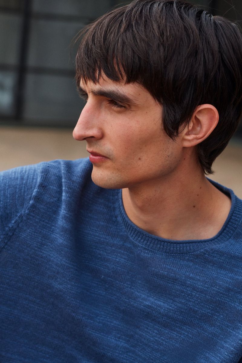 Close-up portrait of a young adult male with dark hair, wearing a textured blue sweater, gazing off-camera in an outdoor setting.