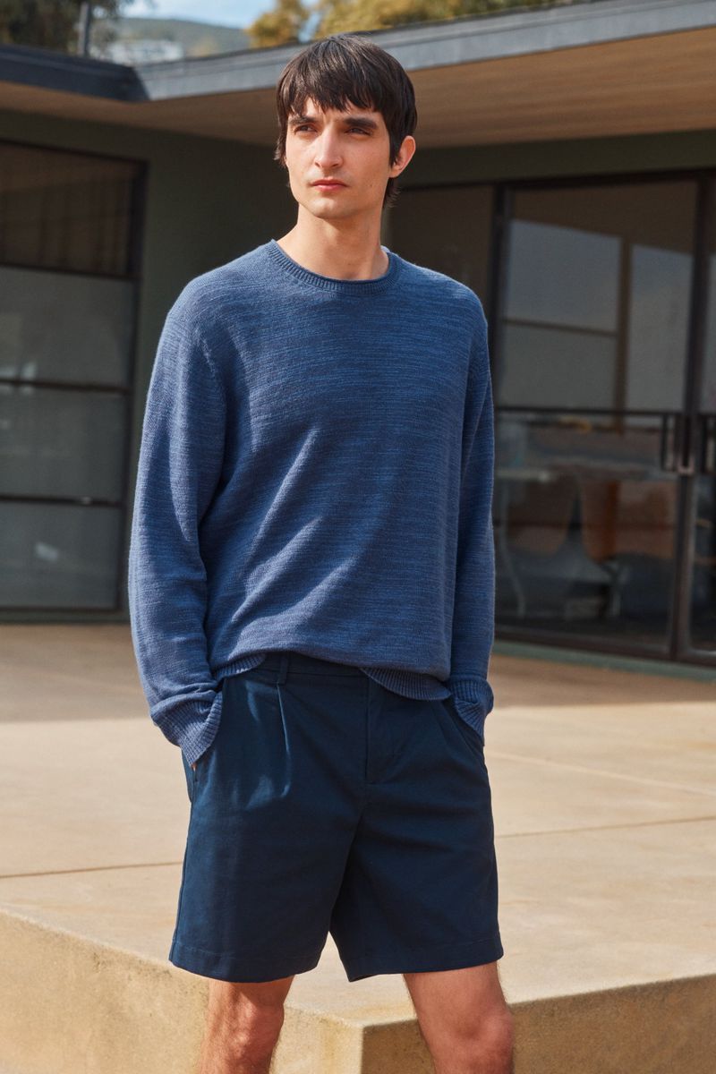 A young adult male in a blue sweater and navy shorts stands outdoors in natural light with a modern building in the background.