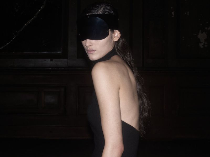 Portrait of a slender adult woman wearing a black dress and a large black blindfold in a dimly lit, dark interior setting.
