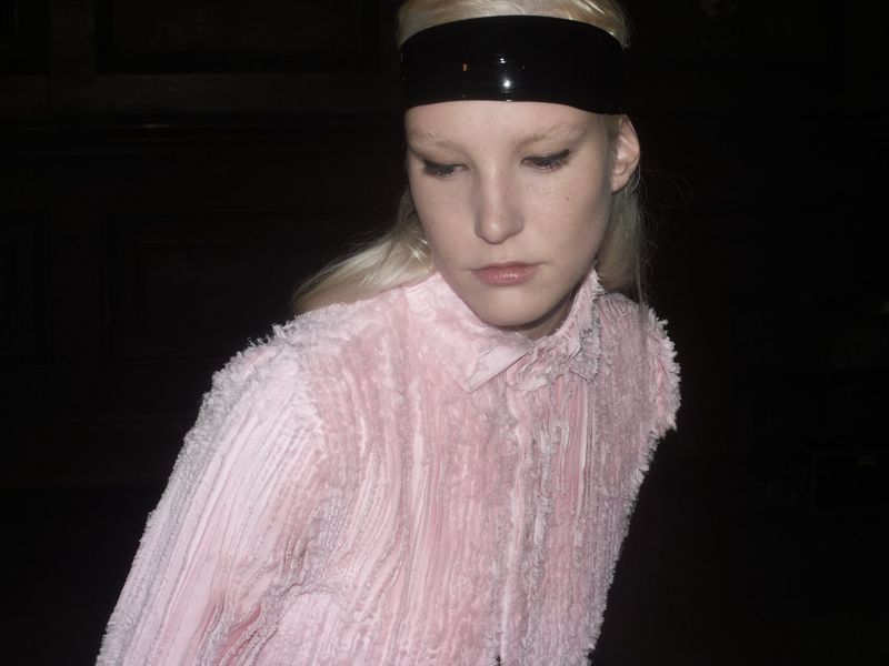 Blonde model with a glossy black headband wears a textured pink button-down shirt against a dark background in a dimly lit indoor space.
