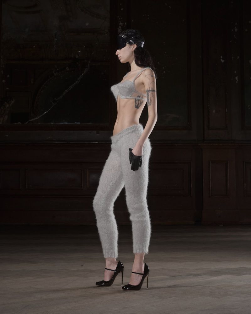 A woman stands in profile, wearing a bra top, fuzzy sweatpants, gloves, and high heels in a room with peeling wallpaper.