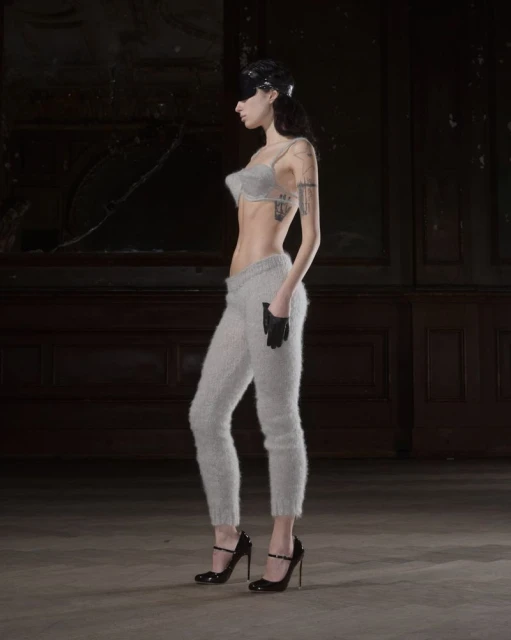 A woman stands in profile, wearing a bra top, fuzzy sweatpants, gloves, and high heels in a room with peeling wallpaper.