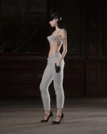 A woman stands in profile, wearing a bra top, fuzzy sweatpants, gloves, and high heels in a room with peeling wallpaper.