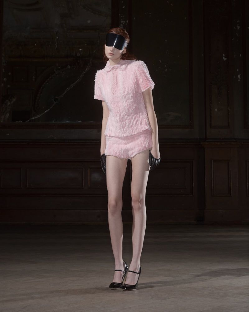 A slender woman in a pink textured skirt suit poses with dark visor, black gloves, and heels against a dark, moody backdrop.