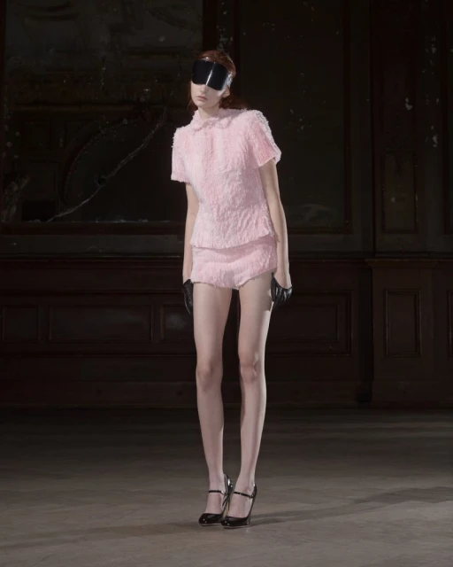 A slender woman in a pink textured skirt suit poses with dark visor, black gloves, and heels against a dark, moody backdrop.