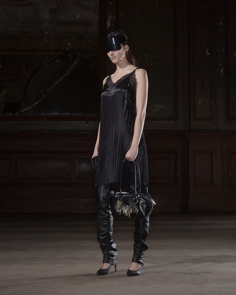 A fashionable woman in a black dress, thigh-high leather boots, and a visor holds a small handbag in a dimly lit interior space.