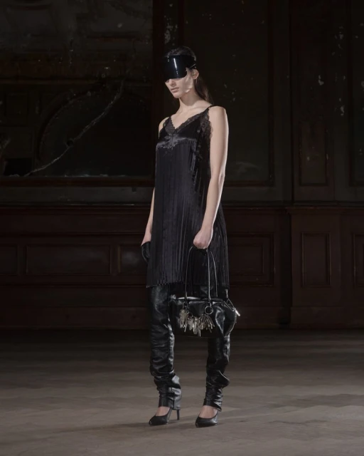A fashionable woman in a black dress, thigh-high leather boots, and a visor holds a small handbag in a dimly lit interior space.