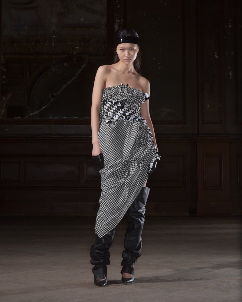 Woman poses in a strapless houndstooth dress and black thigh-high boots against a dark wood paneled wall in an indoor location.