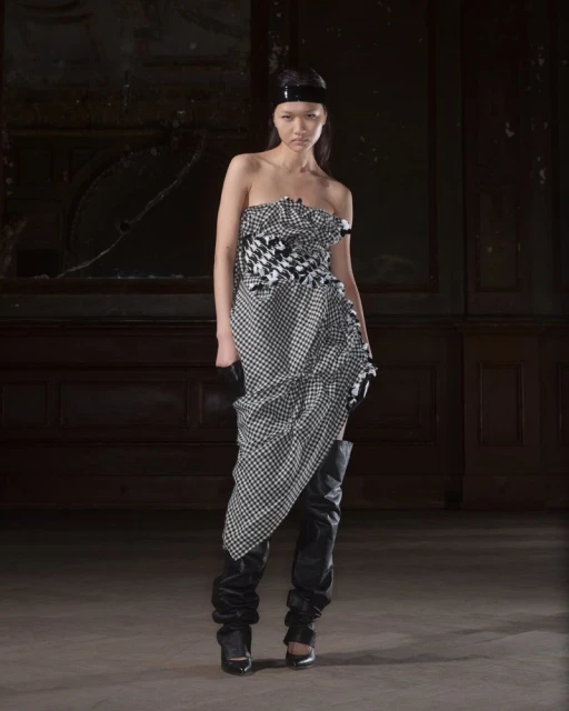 Woman poses in a strapless houndstooth dress and black thigh-high boots against a dark wood paneled wall in an indoor location.