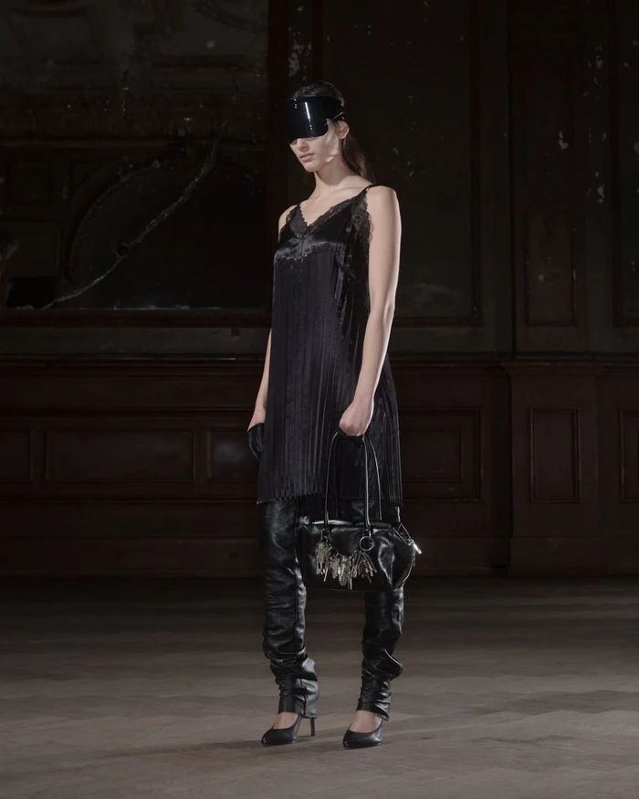 Model standing in a dark interior wearing a black dress, leather boots, visor, and holding a black handbag.