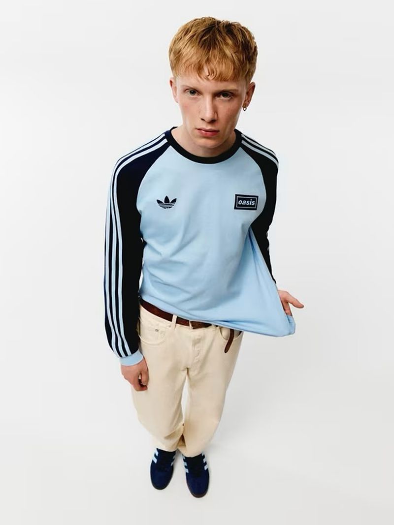 A stylish young adult poses in a light blue long-sleeve Adidas top and beige pants against a bright white backdrop, offering a high-angle view.