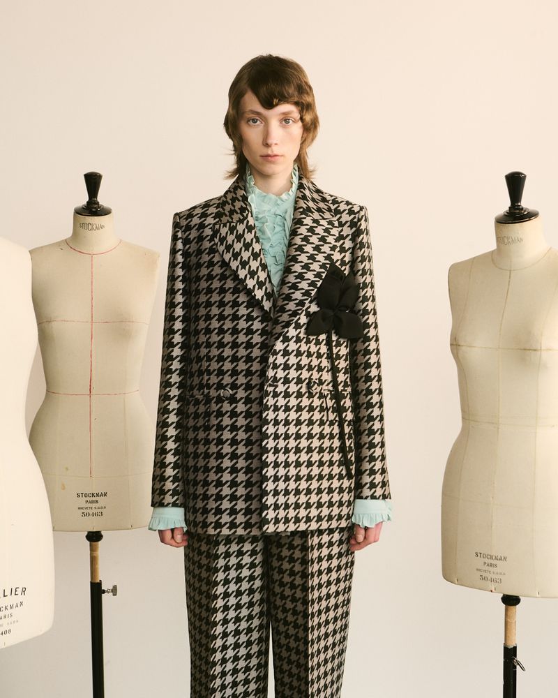 A person in a houndstooth suit stands in a light studio with dress forms. The suit has a black floral detail on the jacket lapel.