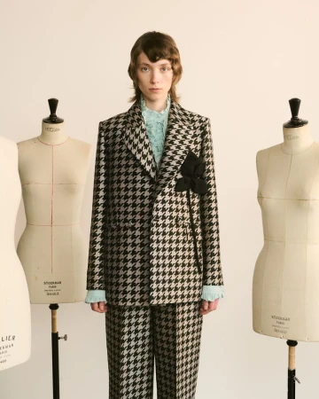 A person in a houndstooth suit stands in a light studio with dress forms. The suit has a black floral detail on the jacket lapel.