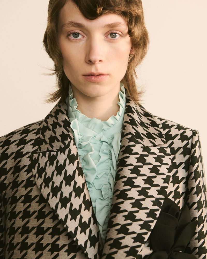 Fashion portrait of a woman in a houndstooth blazer and ruffled blouse, posing indoors with a neutral backdrop, showcasing style and design.