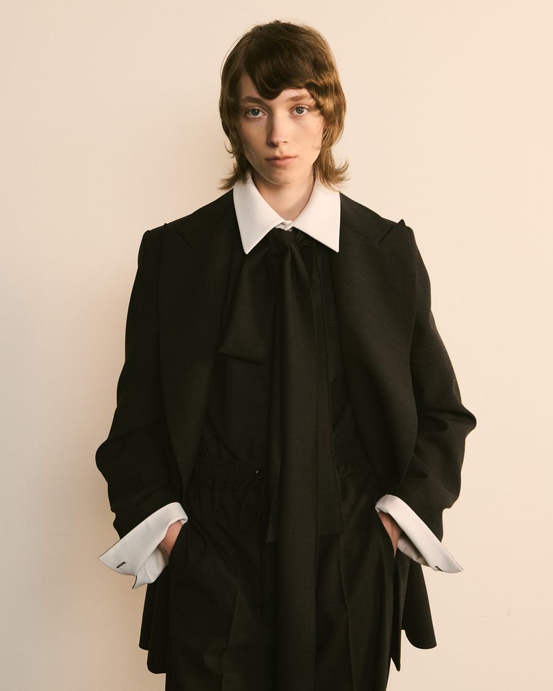 A singular adult model with long brown hair poses indoors wearing a black suit with a white collared shirt and black bow tie.